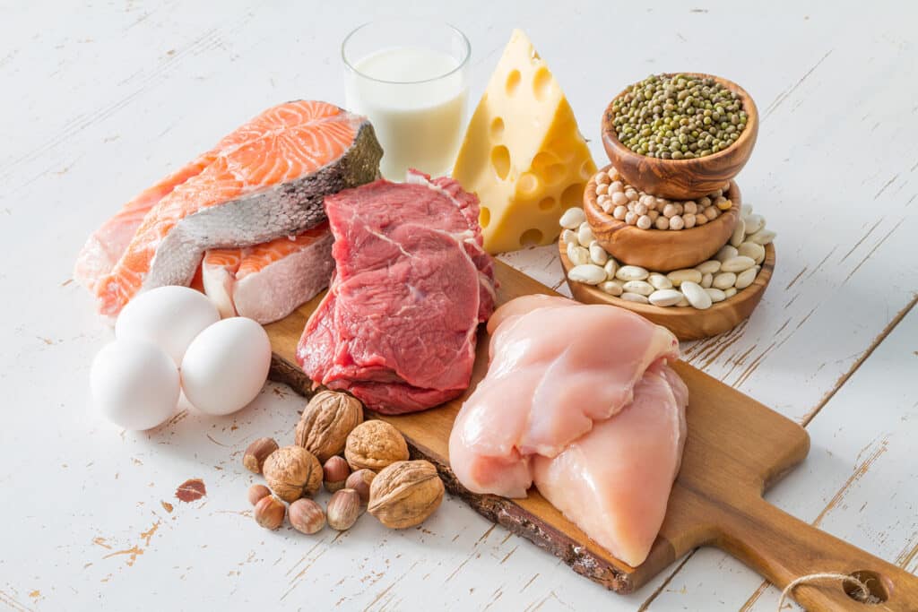 A cutting board holds fresh chicken, beef, and fish. Other proteins such as eggs, cheese, legumes, nuts, and a glass of milk are placed around the cutting board.