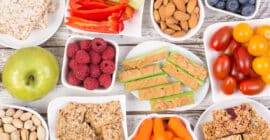 An overhead view of a table with various plates of healthy snacks: berries, granola bars, nuts, and vegetable sticks.