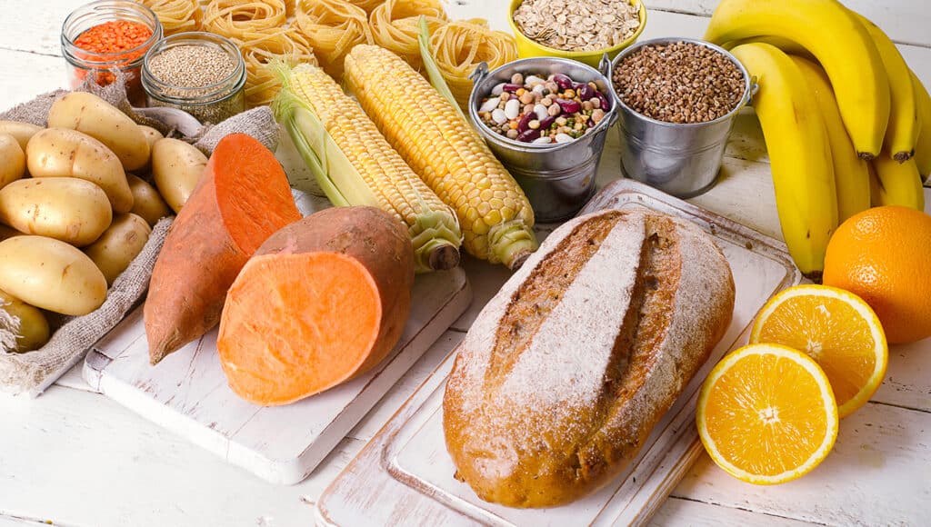 A spread of grains and fruit such as bread, seets, corn, pasta, oranges and bananas. They sit on a table ready for preparation.