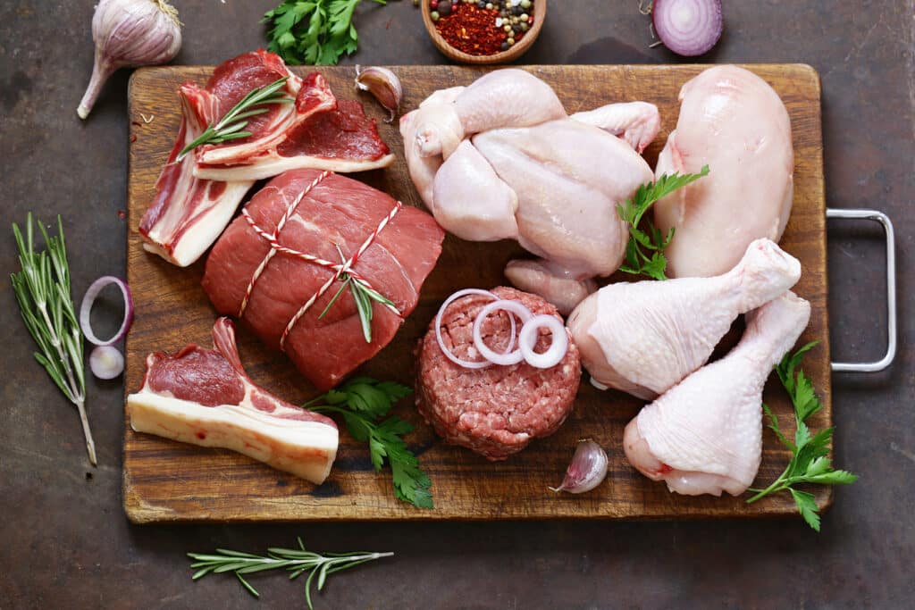 A spread of raw meats such as a whole chicken, chicken breast and legs, ground beef, a roast, etc. sit on a cutting board ready for preparation. Garnish like thyme, parsley and garlic surround the cutting board.