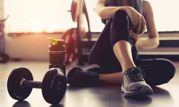 A person sits on a gym floor. Their wrist rests on their bent knee. They wear yoga pants and running shoes. Next to them sits a water bottle and a dumbbell weight.