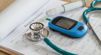 A stethoscope and 2 other medical tools sit on top of a clipboard with a paper titled "Blood Glucose Testing Record".