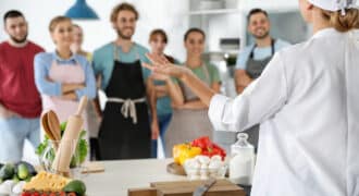 A chef stands with their back to the camera. They address a group of smiling people. Various fresh vegetables and cooking tools sit on a table in front of the chef.