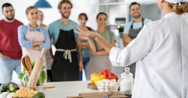 A chef stands with their back to the camera. They address a group of smiling people. Various fresh vegetables and cooking tools sit on a table in front of the chef.