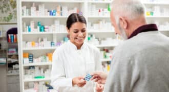 A pharmacist smiles and hands a patient their medication. Shelves of different medications are in the background.
