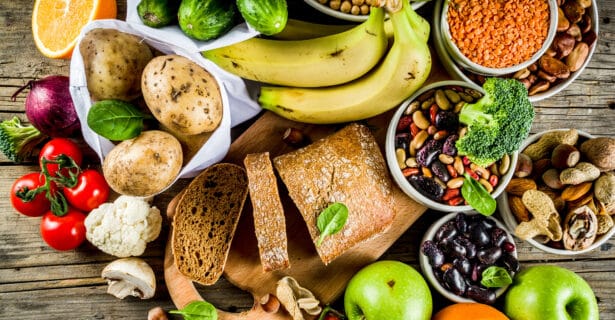 An overhead view of various whole fresh foods in bowls such as legumes, cucumbers, potatoes, apples, and bread sits on a cutting board on the same table.