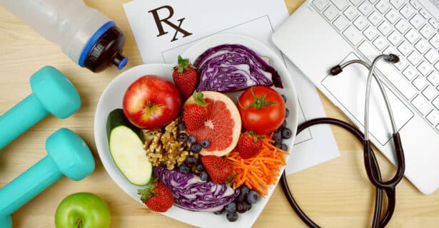 A bowl in the shape of a heart holds fresh vegetables, fruit, and nuts. Under the bowl is a paper with "RX" on the top. A stethoscope and laptop are on the right and weights and a water bottle are on the left.