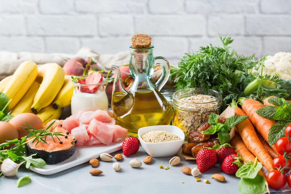 A spread of fresh produce on a table includes carrots, strawberries, oats, nuts, fish, bananas and a jar of oil.