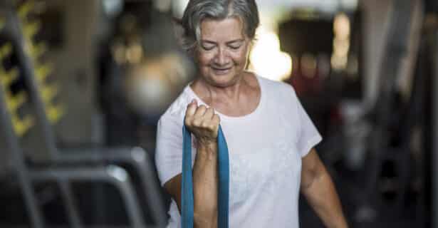 An older woman smiles while she pulls up on a resistance band. She has short grey hair and wears a white t-shirt.