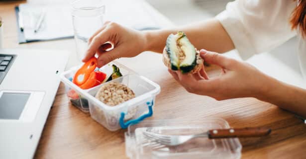 An open container next to a laptop holds vegetables and a rice cake. A hand picks up a pepper. The other hand holds a rice cake with avocado on it.