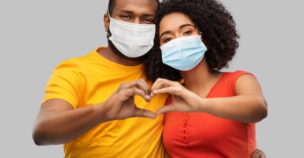 A couple stand next to each other with their heads touching. They bring their hands together to create the shape of a heart. Both are wearing medical masks.