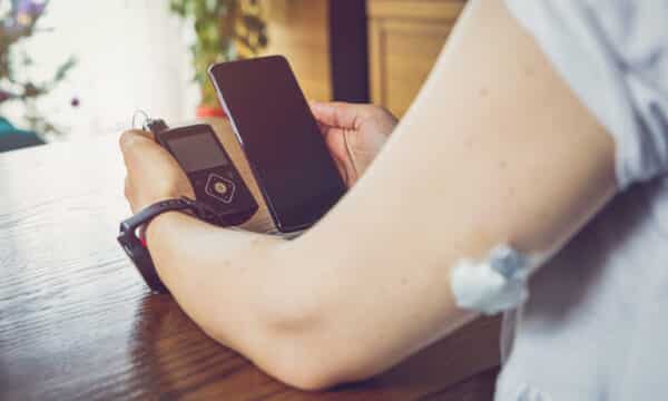 A person holds a glucometer in one hand and their smart phone in the other hand. They have a small monitor on their arm.