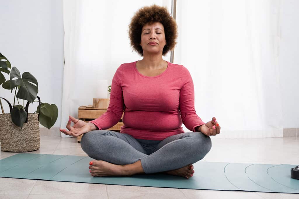 A person sits on a yoga mat with eyes closed while meditating. Her legs are crossed and hands rest on her knees. She wears work out clothing.