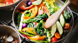 A pan full of fresh vegetable cooks on a stove. A wooden spoon stirs the broccolini, zucchini, tomatoes, peppers, and onions.