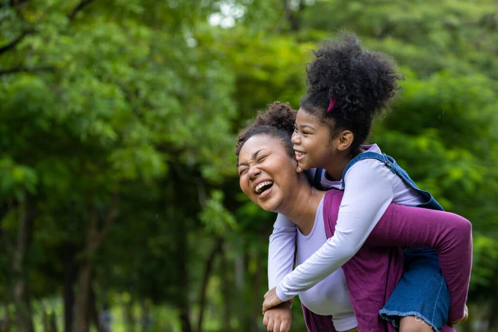 A mother gives her daughter a piggy back ride while they both laugh. Both have curly black hair that is tied up.