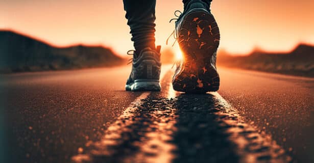 A low view of a road while a person in running shoes walks in the middle of it at sunset.