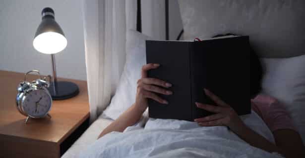 A person reads a book in bed while under the covers. The book covers their face. A clock and lamp sit on the table next to the bed.
