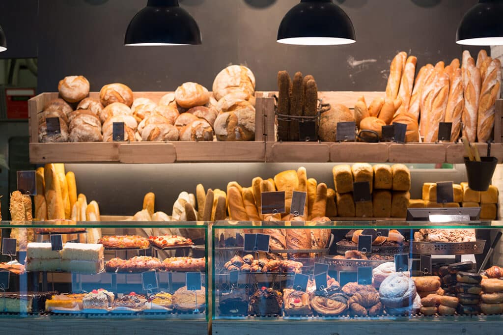 Tall wooden shelves and glass cases in a bakery are full of different types of breads. Square loafs, baguettes, round loafs and more.