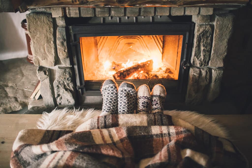 2 pairs of feet that wear fuzzy socks peek out from beneath a warm blanket. They are in front of a lit fireplace.