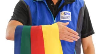 A person wears a UHN vest and has 4 flags draped over their arm in the colours: yellow, red, green, and blue.