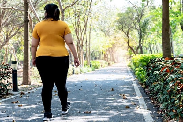 A person walks along a paved path. They wear a yellow shirt and black leggings. Green bushes and trees line either side of the path.